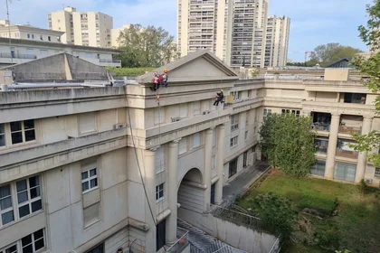 Deux cordistes en façade d'un bâtiment ancien à fronton et arcades